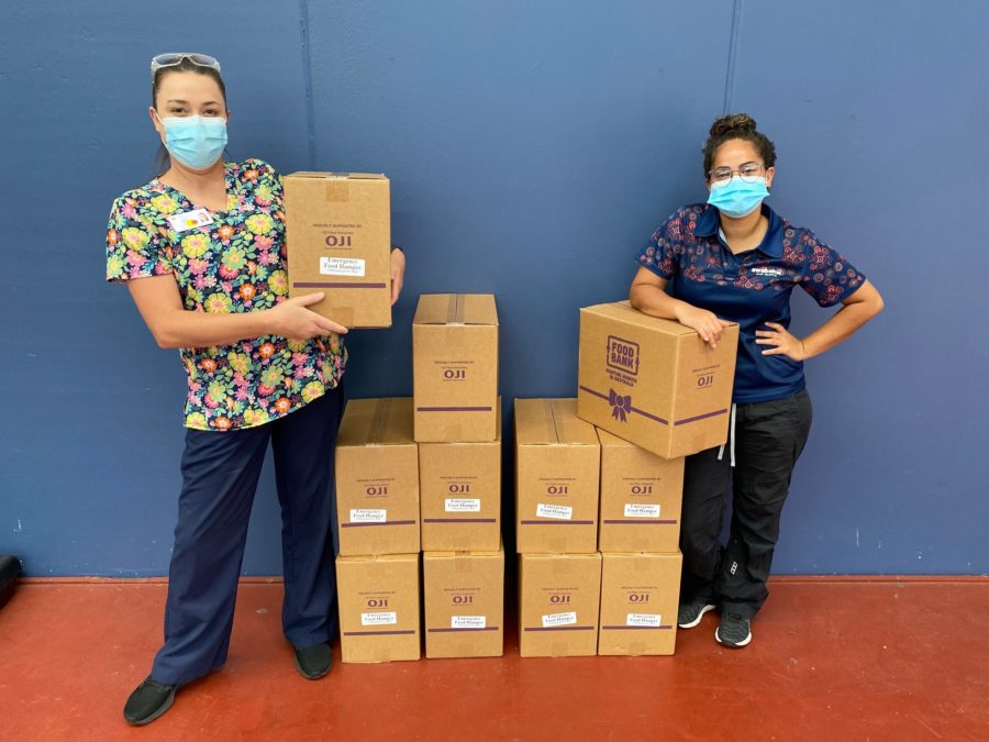 Two healthcare workers wearing face masks stand with stacks of "Emergency Food Hamper" boxes donated by Foodbank and OJI against a blue wall.