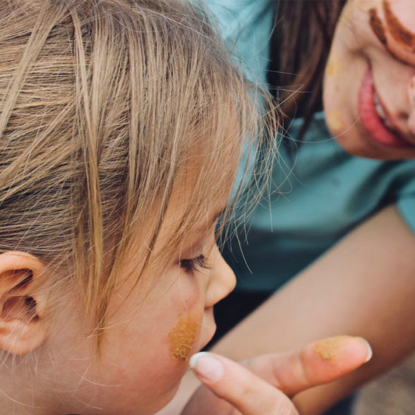 A close-up of a young girl having yellow ochre applied to her cheek by an adult during a traditional cultural activity.