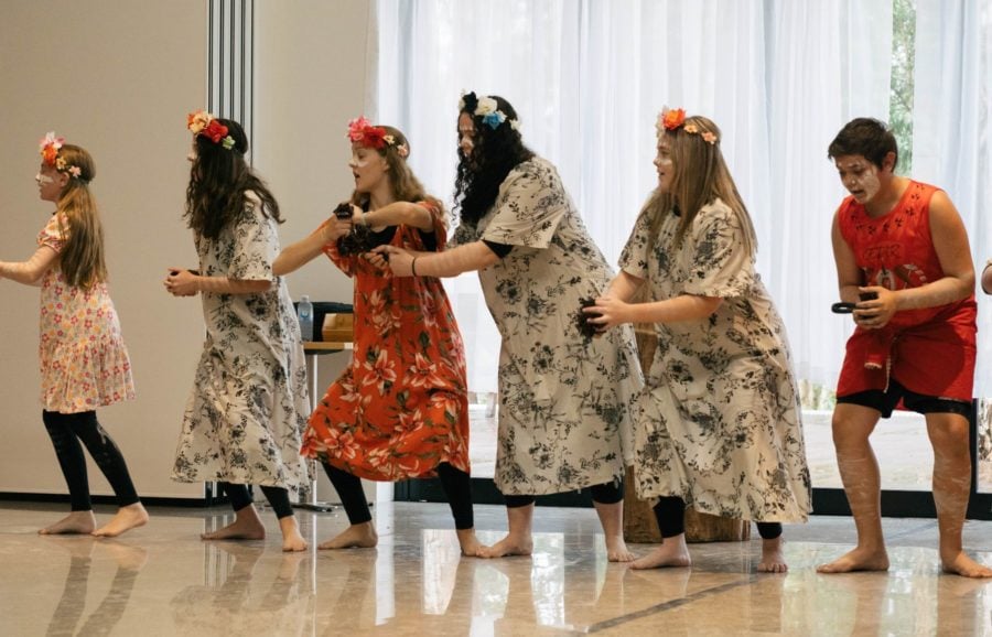 A group of young Aboriginal dancers in floral dresses and headpieces, with white ochre on their faces, perform a traditional dance on a polished indoor floor.