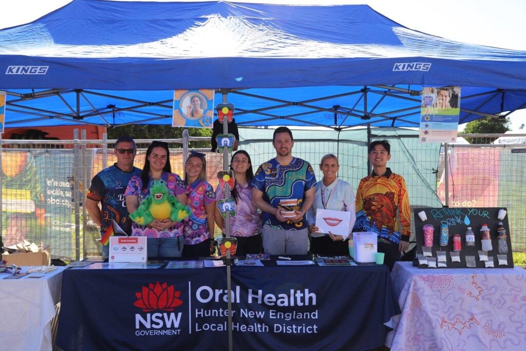 A group of health professionals standing behind a blue NSW Government Oral Health display booth at an outdoor community event
